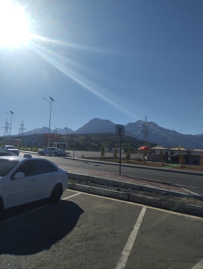 The image depicts an outdoor scene on a sunny day, likely in Dardar, Uzbekistan, with a clear blue sky and bright sunbeams. In the foreground, a white sedan, identified as a taxi by the "TAXI" sign on its roof, is parked in a lot. Its shadow stretches across the asphalt. Adjacent to the parking lot is a paved road with a sidewalk. Beyond the road, there are several structures, including a small kiosk, a covered seating area with red and beige umbrellas, and some buildings in the background. Towering electrical pylons and distant mountains form the backdrop of the scene. The landscape is arid, with sparse vegetation on the lower slopes of the mountains. The overall impression is one of a roadside location with amenities, set against a dramatic mountainous horizon.