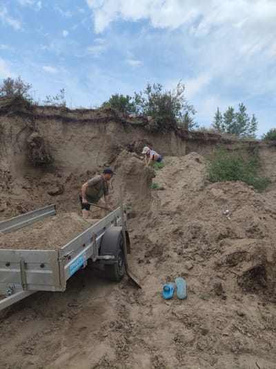In the city of Bel'tirskiy, Russia, a man wearing a dark t-shirt, shorts, and a baseball cap is shoveling dirt into a trailer. A child wearing a light-colored t-shirt and shorts is playing on a sandy incline. The environment is a construction site with a large mound of dirt and a trailer. The sky is blue with scattered clouds. There are also some blue flip-flops lying on the ground.