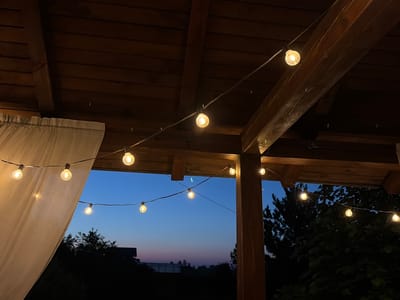 The image shows a string of round, incandescent lights strung under a wooden roof structure. The sky in the background is a deep blue, transitioning to lighter shades of pink and purple near the horizon, indicating dusk or dawn. Silhouetted trees and a structure that appears to be a house are visible against the twilight sky. A white curtain hangs on the left side of the image. The lighting suggests an outdoor or semi-outdoor setting, possibly a patio or porch. There are no people or animals visible in the image. The scene is set in Leninsk-Kuznetskiy, Russia.