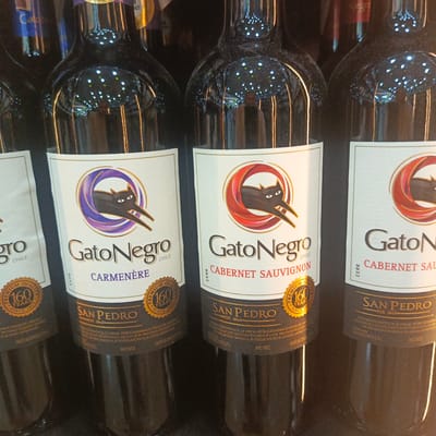 The image displays a close-up view of several bottles of GatoNegro wine. The bottles are dark glass and appear to be in a retail setting, possibly a supermarket or liquor store. The labels are white with a prominent black cat logo. Two of the visible labels clearly indicate "GatoNegro" with "CARMENERE" and "CABERNET SAUVIGNON" below it, along with "SAN PEDRO EST. 1865". The lighting suggests an indoor environment with reflections on the glass from overhead lights. There are no people visible in the image. The image does not provide any specific information about the location being in the city of Los Teques, Venezuela.