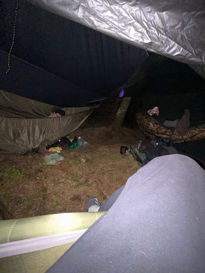 The image shows a forest campsite at night in Macclesfield Forest, United Kingdom. Two people are lying in hammocks, with a third person sitting on a green sleeping mat. The hammocks are suspended between trees, and a tarp is stretched over them for shelter. The ground is covered in brown leaves and twigs. Scattered on the ground are various camping items, including shoes, a plastic bag, and a metal cup. The photographer is situated at the bottom of the image and is looking towards the hammocks. The person sitting on the mat has their back turned to the camera. A purple object is hanging from a branch above. The image was taken at night, and the lighting is dim. The image was likely taken using a low light camera setting. The image shows a small, intimate camping space. There is no evidence of a campfire or any other light source besides the faint glow of a head lamp. The photographer is likely documenting their campsite for their own reference.