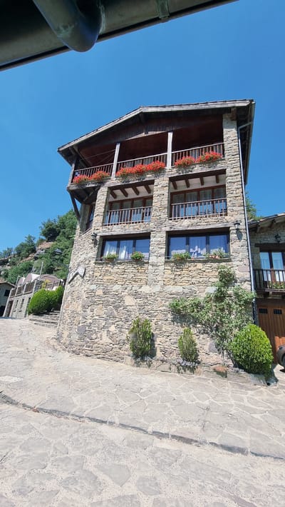 The image shows a traditional stone building in Rupit i Pruit, Spain, under a clear blue sky. The building features a wooden balcony on the upper level adorned with red flowers in planters. Below the balcony, there are two windows on the second floor and two on the first floor, all with wooden frames and visible curtains. The facade of the building is constructed from rough-hewn stones. To the left, a cobbled pathway leads up to another part of the structure, with lush green trees and vegetation on a hillside. To the right, a portion of an adjacent building with a wooden door is visible. Several small evergreen shrubs are planted in front of the main building.