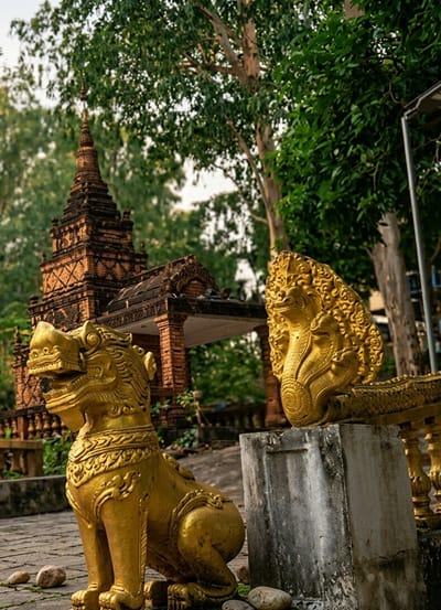 In Sihanoukville, Cambodia, a golden lion statue sits on a stone pathway, facing left with its mouth agape and mane detailed with intricate carvings. Behind it, a multi-tiered brick structure with a pointed roof rises, adorned with decorative brickwork. To the right of the lion, another golden statue, this one a multi-headed serpent, rests on a stone pedestal. Lush green trees with tall trunks form a backdrop, their leaves creating a dappled canopy overhead. The scene is bathed in soft, natural light, suggesting it might be morning or late afternoon.