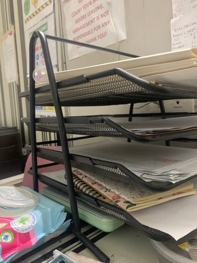 The image shows a black metal desk organizer with multiple trays filled with papers and notebooks. In the foreground, a package of baby wipes with a blue and pink design is visible. The background consists of a wall with posters and a bulletin board. One poster has text that reads "COUNT YOUR MON..." and "BEFORE LEAVING. MANAGEMENT IS NOT RESPONSIBLE FOR ANY LOS... THANK YOU!". Another piece of paper has handwritten text indicating "ELECTRICIAN", "BASIC =", "SSS", "ADMT =", and "PHILHEALTH =". The overall scene suggests an office or workspace environment. The location is identified as Angeles City, Philippines. There are no people visible in the image.
