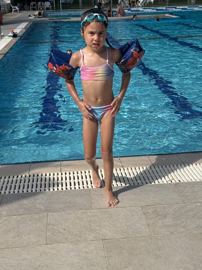 A young girl wearing goggles and arm floaties is standing at the edge of a swimming pool. She is wearing a colorful, striped swimsuit and has her right hand on her hip. The pool water is a bright blue with darker blue racing lanes visible. Next to the pool is a tiled area with a drainage grate. The scene suggests a sunny day at a swimming pool, possibly in a recreational area or a hotel in Beit Yitzhak-Sha'ar Hefer, Israel. The girl appears to be looking at the camera with a neutral expression. Other swimmers and lounge chairs are visible in the background, indicating a public or shared pool facility.