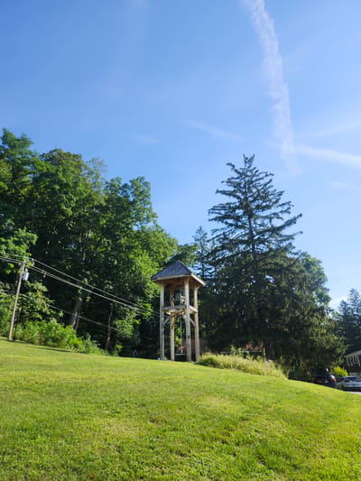 A low-angle, full outdoor shot captures a clear blue sky with a wispy contrail high above. The foreground is dominated by a lush, green grassy hill. In the mid-ground, a wooden bell tower stands prominently on the hill. The tower is constructed of light-colored wood and has a peaked roof. A bell is visible within the tower. Behind the bell tower, a dense, dark green forest of deciduous trees fills the background. To the left of the bell tower, a utility pole with electrical wires stretches across the frame. To the far right of the image, a residential building and two parked cars are partially visible on a paved surface. The overall scene depicts a tranquil, rural or suburban setting.