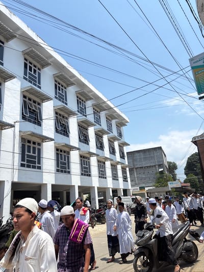 The image captures a bustling street scene in Mideuenjok, Indonesia, on a bright, sunny day, viewed from a low angle looking upwards. The clear blue sky is crisscrossed by numerous black electrical wires.
On the left, a prominent multi-story white building features many rectangular windows, some of which are open, and ornate, patterned awnings or small balconies on each level. To the right, a line of men and boys, predominantly dressed in white Islamic garments such as kurtas/thawbs and kufis/pecis, are either walking or standing alongside parked motorcycles. Some individuals wear patterned sarongs or shirts. They appear to be engaged in a gathering or procession.
In the background, behind the white building, a concrete, possibly unfinished, structure is visible. Further right, along the street, other commercial buildings are present. Several motorcycles (scooters) are parked on the side of the road, with some of the men either standing next to them or seated on them.
Visible text includes "BSI SMART FOTOC FOTO HIDAYA" on a green sign that also lists services like "MELAYANI Transfer, Setor T, Bayar, Toker, Pulsa, Data, Pak, Tog." Other signs display "BSI ATM" and "atm." A motorcycle license plate reads "BL5055 ZBB."