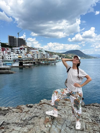 A woman with braided brown hair, wearing a light-colored t-shirt, patterned pants, a white baseball cap, and white Crocs, poses on a rocky outcrop overlooking the Black Sea in Kiparisnoe, Ukraine. She has one hand behind her head and the other on her hip, with her right leg propped up. The sea is a deep blue, and in the background, a town with white buildings stretches along the coastline, with a construction site featuring a crane visible on a hillside. A large, dark green mountain rises in the distance. The sky is filled with dramatic white and gray clouds.