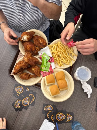 A table laden with fast food, including fried chicken, french fries, and rolls, arranged on disposable plates and a brown tray. Condiments are scattered among the food. A partly consumed beverage, a water bottle, and several Harry Potter themed playing cards are on the table. Two people are visible; one opening a packet of condiment.