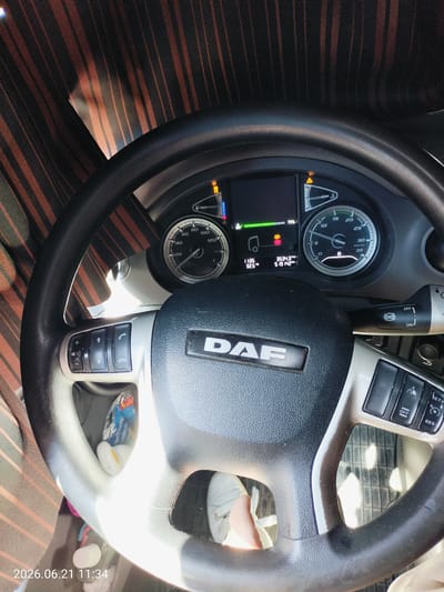 This is a photograph taken inside a DAF truck's cabin. The steering wheel is prominently displayed, with various controls for the infotainment system and cruise control. The dashboard shows a digital display with information such as speed, fuel level, and time. There's also a physical tachometer and speedometer. Sunlight streams into the cabin, illuminating the interior. A pair of white sneakers are visible on the driver's feet. A date and time stamp in the bottom left corner reads "2026.06.21 11:34". There is no visual information in the image to suggest the location is Gorizia, Italy.