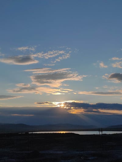 This is a vertical, eye-level, outdoor, medium-shot photograph of a sunset over a body of water. The sun is low on the horizon, partially obscured by dark clouds, but its rays are visible, streaming downwards in shafts of light. The sky is a gradient of blue and orange, with scattered white and grey clouds. In the middle ground, a large body of water reflects the warm colors of the sky. Beyond the water, distant, dark hills form a silhouetted landscape. The foreground is dark and indistinct, suggesting a field or barren land, with a dark fence post visible on the right side. The overall mood is peaceful and serene, capturing the beauty of the natural landscape during sunset. The location is identified as Mayskiy, Russia.