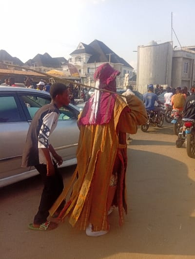 The image depicts a bustling street scene in Ozuri, Nigeria, under clear daytime conditions. In the immediate foreground, facing away from the viewer, stands an individual adorned in elaborate traditional attire, consisting of a flowing, earth-toned (gold/tan/orange) outer garment with maroon/red fabric details on the back and a matching maroon head covering. This individual holds a slender wooden staff vertically, extending above their head. To the left of this person, a young male, possibly a teenager, walks in profile beside a silver sedan. He wears a black long-sleeved shirt with white patterned sleeves, black trousers, and green patterned flip-flops, looking towards the right side of the frame.
The midground features a silver sedan on the left, with numerous individuals and multiple motorcycles (Okadas) further down the unpaved road. Riders and passengers are visible on the motorcycles, contributing to a sense of activity. In the background, prominent buildings include a multi-story white structure with a dark, multi-peaked roof, and to its right, a large cylindrical concrete structure (potentially a water tower or silo) adjacent to another building. The ground is an unpaved surface, and the bright sunlight casts distinct shadows. Utility poles and wires are visible against the clear sky.