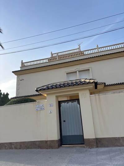 The image shows the facade of a villa named "VILLA ALVERA" with the number 21 on a sunny day. The villa is painted in a light yellow color, and has a tiled roof. A security camera is visible on the upper part of the wall. A paved walkway runs in front of the villa. The sky is clear and blue, with some power lines crossing it. In the background, there are trees and a balcony on the roof of the villa with decorative railings.
