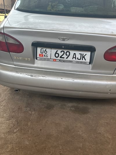 A rear view of a silver Daewoo car is captured in a dimly lit environment, likely a garage or parking area, with a concrete floor. The car's license plate, 06 KG 629 AJK, is clearly visible and features the Kyrgyzstan flag and text. The bumper shows signs of wear and tear, with peeling paint. An exhaust pipe is visible at the bottom left of the image. The background is blurred, obscuring most details of the surroundings.