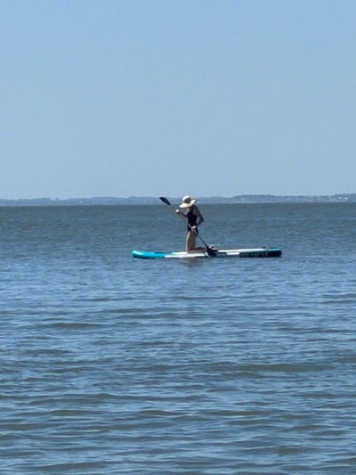 A lone individual is paddleboarding on a calm body of water. The person is wearing a dark swimsuit and a wide-brimmed hat, kneeling on the paddleboard and holding a paddle. The paddleboard is white with blue accents. The water is a deep blue with gentle ripples, and the sky above is a clear, pale blue. In the distance, a hazy, landmass is visible along the horizon. The scene appears to be set in an open, natural environment.