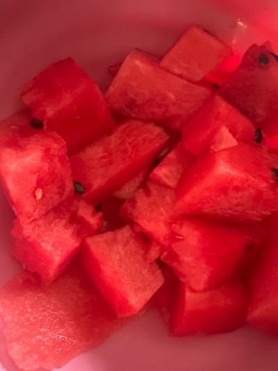The image is a close-up, overhead shot of several chopped pieces of red watermelon, some with black seeds, in a pink bowl. The lighting is warm and slightly reddish, possibly due to artificial light. There are no people or any identifiable landmarks of the city of Konstantinovo, Russia present in the image.