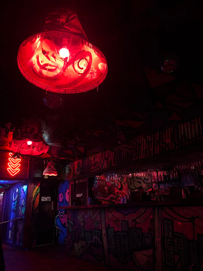 This image depicts the interior of a dimly lit bar or entertainment venue located in West Lancashire, United Kingdom, characterized by a predominantly red light ambiance. The ceiling features a dark, patterned surface with geometric shapes and sections of corrugated metal. A large, fringed lampshade, emitting a bright red light from a central bulb, hangs prominently in the foreground, displaying swirling patterns on its surface. Another smaller, similarly styled lampshade is visible further back. A small disco ball is mounted on the ceiling.

The walls are adorned with extensive, multi-colored graffiti-style artwork, including abstract designs, figures, and urban landscape motifs. A bar counter, also heavily decorated with graffiti depicting a cityscape, extends across the midground. Behind the bar, shelves hold bottles and glassware, with some glasses inverted above the counter. To the left, a black door marked "GENTS" in white capital letters indicates a restroom. Adjacent to this door, a red neon sign shaped like three downward-pointing chevrons illuminates the area. No individuals are visible within the scene.