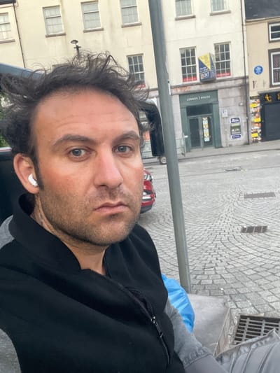 A selfie of a man with short, dark, unkempt hair and stubble. He is wearing a black jacket over a gray shirt and has a white wireless earbud in his left ear. The background shows a city street with light-colored stone pavement and several buildings. One building has multiple windows and a sign indicating "Lennox House." The image appears to have been taken in the city of Armagh, United Kingdom, based on the architectural style and street layout. There are also some signs and advertisements visible on the buildings in the background.