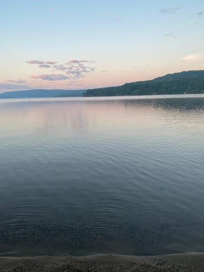 The image depicts a serene lake scene at sunrise or sunset. The sky is a soft gradient of blue and pink, with scattered wispy clouds. A forested hill forms the horizon line across the lake. The water is calm with gentle ripples, reflecting the colors of the sky. In the foreground, the edge of a pebble beach is visible. The scene appears to be located on the shores of Lac Simon in Duhamel, Quebec, Canada, judging by the characteristic landscape and the likely geographical context of the prompt.