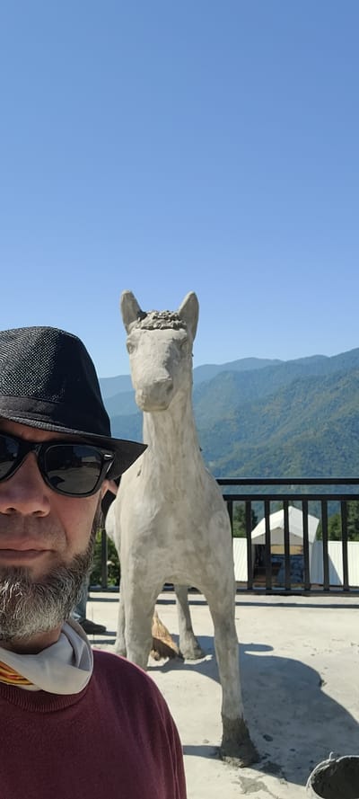 A man wearing a black hat, sunglasses, and a maroon sweater is taking a selfie in front of a concrete horse sculpture. The man has a grey beard and a light-colored scarf around his neck. The sculpture is unfinished, with visible concrete on its legs. In the background, there are lush green mountains and a clear blue sky. A black railing with white structures behind it is visible between the sculpture and the mountains. The scene appears to be outdoors, possibly on a rooftop or balcony.