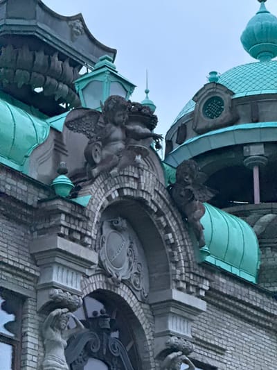 The image depicts the facade of an ornate building with prominent architectural features. The primary color palette consists of muted grays, blues, and teal, suggesting an overcast sky or the aged patina of the materials.
In the foreground, the building's exterior is constructed of light gray bricks, laid in a regular pattern. Above the brickwork, several decorative elements are visible. To the left, a sculpted female figure is partially visible, her arms raised above her head, carved from a light-colored stone. She is positioned above an arched window opening.
Further up, a stone archway frames a decorative crest or emblem. Above this arch, on the roofline, are several winged putti sculptures. One putto is seated, holding what appears to be a small object or garland. Another putto is perched precariously on the edge, with outstretched arms. These sculptures are dark in color, contrasting with the lighter stone and brick.
The building's roofline is adorned with several green, dome-shaped structures, likely made of copper or a similar material, which have oxidized to a teal hue. These domes are tiled and feature finials at their apex. Intricate metalwork, possibly decorative railings or cresting, is visible along the roof edge. Above these, a dark, tiered structure with carved faces and repeating motifs can be seen, further emphasizing the building's elaborate design.
The overall impression is of a historic, possibly institutional or religious, building with a rich architectural history. The image does not provide specific clues to locate it in Valday, Russia.