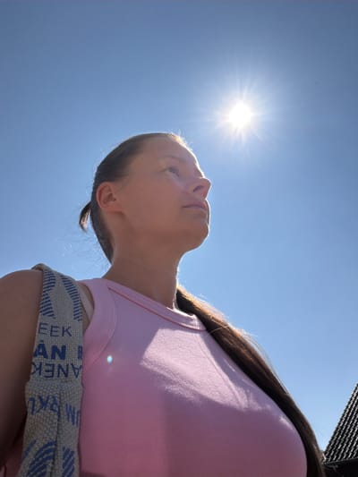 A low-angle, medium shot captures a young woman from the chest up, looking upwards towards a bright sun against a clear blue sky. She has long, dark brown hair pulled back into a ponytail. The woman is wearing a pink tank top, and a bag with blue and white stripes is slung over her left shoulder. The bag has some text visible on it in blue lettering. A dark roofline is visible in the lower right corner of the frame, suggesting an outdoor urban or suburban setting. The image was taken in Melick, Netherlands.