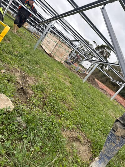 The image depicts a construction site for a solar farm on Baldurgan Hill, Ireland, under an overcast sky. Numerous rows of newly installed metallic support structures, likely for solar panels, extend across the scene. The foreground consists of uneven ground with patches of green grass, clover, and disturbed earth, along with scattered small rocks.
In the upper-left, a male construction worker is visible, wearing a dark jacket with orange accents, dark trousers, a blue hard hat, yellow gloves, and light-colored work boots. He is leaning forward, seemingly interacting with or inspecting a horizontal beam of the solar array framework.
Underneath the solar structures, a large, light-brown wooden crate with green strapping and stenciled markings is situated on the ground. Adjacent to the crate, electrical components are visible, including a white inverter box (possibly Huawei brand) attached to the frame, with bundles of black cables running from it. Further back on the right, an orange safety net or fence runs along the edge of a wooded area with mature trees. The lower-right corner of the image shows the mud-splattered blue wellington boot of a second individual, partially cropped out. A bright yellow L-shaped object is partially visible in the bottom-left corner.