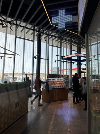 The image displays the interior of a modern commercial establishment in Vorsha, Russia. The space features a high ceiling with exposed dark metal beams and a grid structure, equipped with various linear and spot lighting fixtures. A prominent, large, illuminated LED cross sign hangs from the ceiling in the upper center. Extensive floor-to-ceiling windows on the left and rear reveal an outdoor scene with parked vehicles and a distant red-and-white building, likely a petrol station or retail outlet, beneath an overcast sky.
The interior floor is paved with light-colored tiles, showing reflective patches. In the mid-ground, a woman in a red-brown top, light skirt, dark leggings, and light boots is depicted mid-stride, reaching towards a wooden display counter. This counter has a ribbed base and a glass-topped section showcasing numerous small, individually packaged items. To the right of this counter, a small group of individuals is visible, including a man in a dark jacket and beanie facing right, and another person in a light grey hoodie facing left. A curved overhead sign displaying the text "ЛЭТУАЛЬ" (L'Etoile) is positioned to the right of the LED cross. On the far left, a decorative wooden planter box containing green foliage features a carved burger icon on its side.