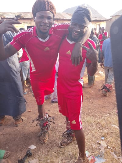 In Vodni, Nigeria, two adult males are positioned prominently in the foreground of an outdoor scene. The male on the left, wearing a brown knitted cap, a red Adidas short-sleeved shirt with white side stripes, and red shorts, smiles directly at the viewer while gesturing a peace sign with his left hand and resting his right arm over the shoulder of the adjacent male. His left lower leg and ankle are adorned with an intricate metallic decorative or symbolic apparatus, secured over a light brown sandal.

The male on the right, sporting a dark fabric head covering, a beard, and a matching red Adidas short-sleeved shirt and red shorts, grins widely, displaying a "rock on" hand gesture with his right hand. His left arm rests over the shoulder of the male on the left. His right lower leg and ankle are similarly adorned with an intricate metallic device, positioned over a patterned brown sandal.

The ground beneath them consists of reddish-brown earth with patches of dry grass and scattered debris, including plastic bags and paper. In the midground and background, other individuals are visible, some also wearing red attire, suggesting a gathering. Behind them, simple structures with corrugated metal roofs and walls are present; one building features faint text that includes "ORPHAN." The scene is illuminated by bright daylight.