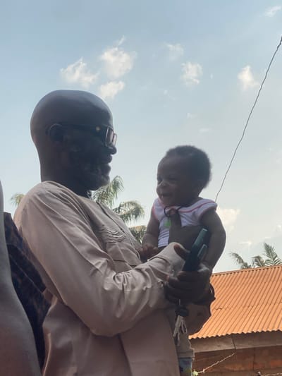 An outdoor scene in Aliade, Nigeria, depicts an adult male holding an infant under a bright, partly cloudy sky. The adult male, with dark skin, a bald head, and a short grey beard, wears dark-framed glasses and a light brown, long-sleeved traditional garment featuring pleated details on the chest. He smiles and looks towards the infant. The infant, with dark skin and short dark hair, wears a white top with pink trim and a thin dark necklace; the infant also smiles, looking towards the adult. The adult male's left hand, which supports the infant, holds a dark-colored object, possibly a mobile phone. A partial view of another individual's shoulder and arm is visible on the left. The background features green foliage, consistent with palm trees, and the orange corrugated metal roof of a building. A thin dark wire diagonally crosses the upper right section of the image.