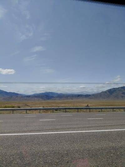 The image captures a highway scene with mountains in the background. The foreground features an asphalt road with white lane markings. A metal guardrail separates the road from a dry, grassy field. Beyond the field, a range of rolling mountains extends across the horizon under a partly cloudy blue sky. The overall impression is one of a rural, open landscape. The scene is likely situated in or around the city of Ынталы, Kazakhstan, a region known for its vast plains and mountainous terrain. No people or specific actions are visible in the frame.