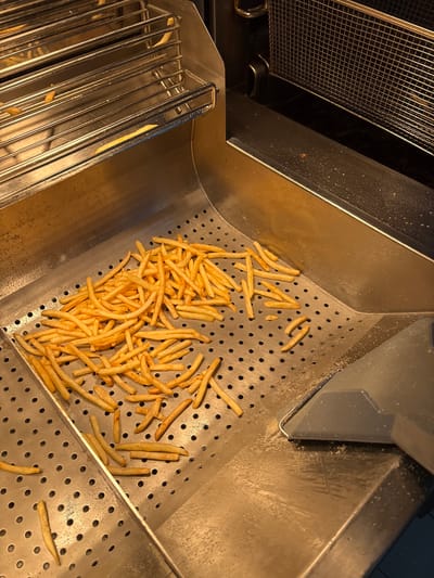 A commercial food service area displays a large quantity of golden-brown, cooked French fries, primarily scattered across a perforated stainless steel warming or holding tray. Above the main collection area, a multi-tiered metal rack, also stainless steel, contains additional fries. To the immediate right, a dark, empty deep fryer basket is partially visible within its housing, indicating proximity to a frying station. Below the fryer basket, the surface is a darker, smooth stainless steel. In the lower right corner, a gray plastic or metal scoop/scraper rests against the perforated tray, with some white crystalline residue, likely salt or seasoning, visible on the adjacent surfaces. The overall environment consists of brushed stainless steel equipment under warm, artificial lighting. The surfaces show some evidence of grease and seasoning from active use. The scene is presented as being in Chuchuligovo, Bulgaria, though no specific visual identifiers within the image confirm this geographical location. No people are visible.
