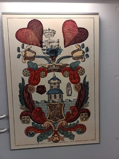 A framed historical illustration is depicted vertically on a light-colored wall surface within an interior setting in Altenburg, Germany. Two silver-toned handles are visible on the left edge, suggesting the display surface is a door or cabinet.
The illustration itself, enclosed in a simple light-colored frame, features a complex heraldic or allegorical design against an off-white background. At the top, two large, textured red heart shapes flank a smaller, faded shield surmounted by a crown. Below this, ornate floral and foliate scrollwork in red, blue, yellow, and green cascades downwards, framing a central scene.
The core of the illustration presents a stylized grey crenellated city wall, behind which stands a prominent, multi-tiered white tower with a blue conical roof. In front of the wall, a shield displays a rampant golden lion on a light field with a dark border. Adjacent to the tower, a dark red rose is positioned on the left and a white open hand symbol on the right. Additional circular floral motifs are integrated into the red scrollwork. The entire composition is outlined by a thin black border. The overall color palette is muted, consisting of reds, blues, greens, yellows, greys, and whites.
