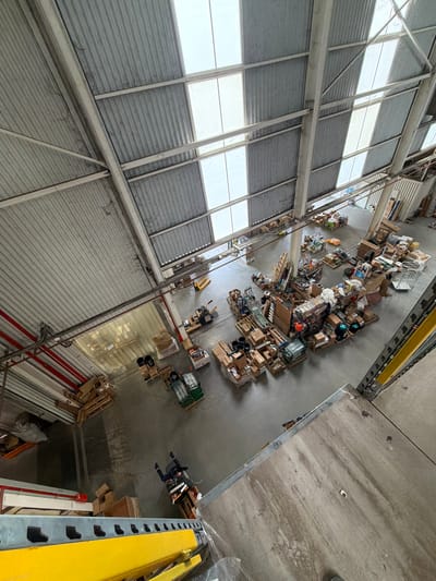 The image depicts an aerial view inside a large industrial building in Stroevo, Bulgaria. The building has a high ceiling with corrugated metal siding and a series of windows. The floor is concrete and appears to be cluttered with various objects, including boxes, pallets, and other materials indicative of warehouse storage. A person is visible on the floor, dressed in dark clothing. Shelving units are visible along the right side of the image.