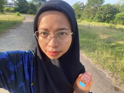 A woman wearing a black hijab and glasses is taking a selfie on a paved road lined with dry grass and trees. She is wearing a dark blue garment with a pattern of blue and black lines and leaf-like shapes. In her right hand, she holds an orange water bottle with a blue lid. The road curves to the left in the background, leading towards a distant building and more trees. The sky is partly cloudy and the sun appears to be shining. The location is identified as Gudang Batu, Indonesia.