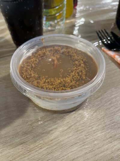 This image displays a multi-layered dessert presented in a transparent, circular plastic container, positioned on a light-colored wood-grain table surface. The dessert consists of a bottom layer of compacted light brown crumbs, an intermediate white creamy layer, and a top layer of smooth, viscous light brown liquid, partially covered with dark brown coarse crumbs. A small, darker, amorphous object is embedded within the top liquid layer. The table surface is covered with a clear, wrinkled plastic film. In the blurred background to the left, a dark beverage with visible carbonation is contained within a clear cup. To the right, a black-handled fork rests beside a folded, orange and white patterned napkin. Other indistinct objects, including a clear plastic bottle, are visible further in the background. No individuals or dynamic actions are present. The image contains no discernible information to confirm the specific location in the city of Kasak, Bulgaria.