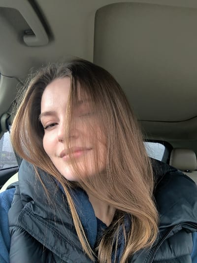 The image displays a headshot of a female subject, approximately 20-30 years old, situated inside the passenger compartment of a vehicle. Her light brown hair is parted on the right, with strands falling across the left side of her face, partially obscuring her left eye. She has light skin, a slightly raised left eyebrow, and a subtle smile. Her eyes appear to be light in color. She is wearing a dark blue or black quilted puffer vest over a lighter blue garment, possibly a hoodie or sweatshirt, with a visible collar. The vehicle's interior features a light-colored headliner and a gray overhead grab handle visible in the upper left. A beige headrest from a rear seat is partially visible in the upper right quadrant, and a dark dashboard or door frame is visible to the left. The scene is illuminated by ambient light, suggesting daytime conditions, with a blurred exterior visible through a window on the left, indicating movement or a soft-focus background. The specific location within the city of Illintsi, Ukraine, is not discernible from the visual information provided.