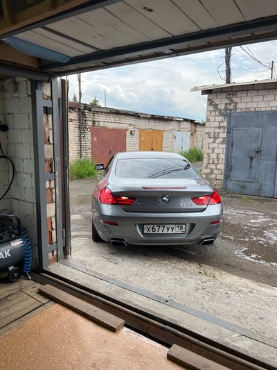 A grey BMW 650i is parked in front of an open garage in a city, possibly Veteran-2, Russia. The car is viewed from the rear, and its license plate reads "X677YY 18 RUS." To the left of the car, an air compressor is visible. The garage has a concrete floor and brick walls. Behind the car are several other garages with metal doors, and the sky is overcast.