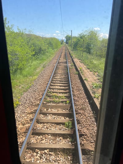 A view from a train window shows a straight section of railroad tracks receding into the distance. The tracks are flanked by ballast stones and vegetation on either side. Overhead power lines run parallel to the tracks. The scene is bathed in daylight under a blue sky. Based on the metadata, this image was captured in Gabarevo, Bulgaria.