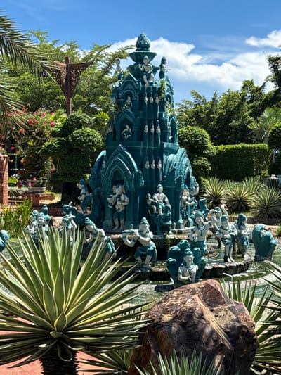 The image shows a multi-tiered turquoise-colored monument adorned with figures, possibly depicting a scene from mythology or local folklore. This structure is situated within a landscaped garden, identifiable by various plant species, including palm trees, agave plants, and other greenery. A large rock sits adjacent to the monument, adding to the natural elements of the setting. The sky is visible in the background, indicating it is daytime. The location is Bang Pu Subdistrict Municipality, Thailand.