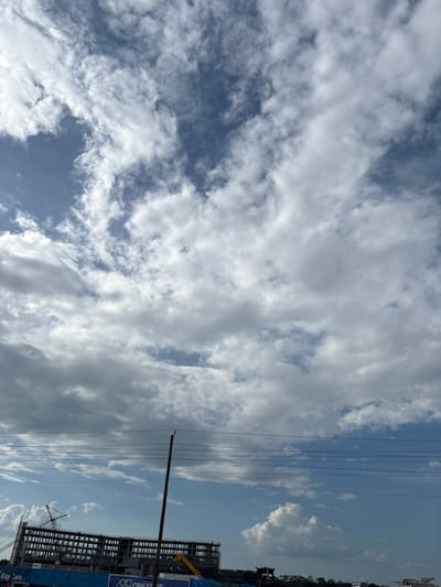 The image captures a construction site in Ellenton, Florida, United States, under a partly cloudy sky. The primary subject is a large, unfinished building structure with multiple floors, its framework exposed, suggesting ongoing construction. Several cranes are visible, one prominently positioned to the left of the building, and another yellow construction vehicle is situated near the base of the structure. Power lines stretch across the middle ground of the image, supported by a utility pole. The background consists of a sky filled with a mix of white, puffy cumulus clouds and darker, more diffused clouds against a blue sky. A blue banner with white lettering is partially visible at the bottom of the building, reading "Bay Care" and "CREATIVE". The scene conveys an industrial and developing urban environment.