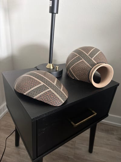 This image presents a thoughtfully arranged indoor scene, showcasing a collection of modern home decor elements. Dominating the right side is a minimalist black bedside table or small cabinet, featuring a single, sleek brass handle on its drawer.

Perched on the table are two distinct ceramic pieces, both adorned with an intricate, repeating geometric pattern. The pattern consists of light pinkish-tan bands that crisscross over a dark background, which is filled with tiny, contrasting dots. One piece is an upright vase or pot with a wide, light-colored rim, while the other is a smaller, oval-shaped decorative item resting horizontally, displaying the same elaborate design.

A modern black floor lamp is positioned to the left, its slender arm extending horizontally across the frame. A small brass ring accents the lamp near where its arm connects to the base. A small, white rectangular label with indistinct black text is visible on the lamp's arm.

The background features a light, textured wall, providing a clean contrast to the darker furnishings. The floor, visible in the lower right, is a light-toned wood or laminate with a subtle grain. A black power cord trails from beneath the table, adding to the domestic setting. The scene appears to be an interior space, possibly a bedroom or living area, in Redondo Beach, United States of America. No specific activity is occurring, and no people are present. The lighting suggests ambient indoor illumination.