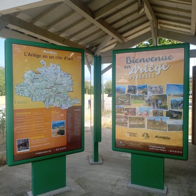 This media file captures a sunny outdoor scene featuring two large informational display boards. The boards are green-framed and mounted on green posts, standing on a concrete surface. The backdrop suggests a public area, possibly a rest stop or tourist information point, with trees and a road visible behind the displays.
The left board, titled "L'Ariège en un clin d'œil," presents a map of the Ariège region of France, along with lists of "Offices de Tourisme" and "Routes Touristiques." There's a small image of a historical building with outdoor seating. The right board, with the title "Bienvenue en Ariège Pyrénées," showcases a collage of photographs depicting various activities and landscapes in the region, including cycling, kayaking, paragliding, and scenic views of villages and natural formations. Text on this board includes "Ariège le Vélo!" and "Grands Sites de Midi-Pyrénées."
The overall atmosphere is bright and inviting, suggesting a pleasant day for outdoor exploration. The presence of detailed maps and photographic highlights indicates that these displays are meant to inform and attract tourists to the Ariège region. The visible text provides website addresses and phone numbers for further information, facilitating planning for visitors. The time of day appears to be daytime, under clear or partly cloudy skies, given the shadows and the brightness of the scene.