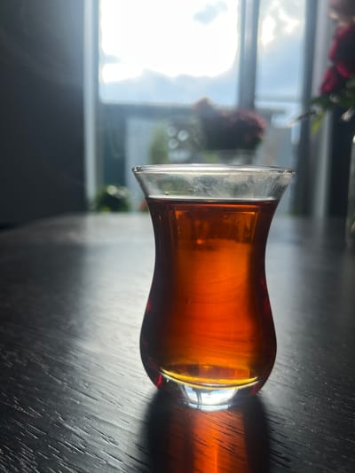 A glass of amber liquid, likely tea, sits on a dark, textured surface, possibly a wooden table. The glass has a curved shape, wider at the bottom and tapering towards the top with a flared rim. The liquid within is clear and luminous, catching the light and casting a shadow on the table. The scene is illuminated by natural light coming from a window in the background, which is bright and slightly overexposed, suggesting it's daytime. Outside the window, blurred elements of greenery and possibly flowers are visible, along with a glimpse of the sky. The overall mood is serene and contemplative, with the play of light and shadow creating a dramatic effect. There is no visible text.