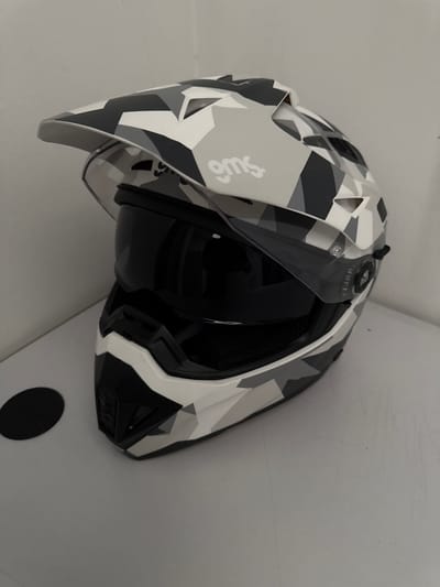 The image shows a close-up, side view of a motocross helmet with a camouflage pattern of white, gray, and black geometric shapes. The helmet is resting against a plain, light-colored wall, possibly in an indoor setting. The visor is partially open, revealing the black interior padding of the helmet. A small, circular, black object is visible on the wall in the lower right corner of the image. There is some scuffing and scratching visible on the wall surface. The helmet appears to be new or well-maintained. The brand name "GMS" is visible in white text on the side of the helmet, near the front. The lighting is even and appears to be artificial, suggesting the photo was taken indoors. No people are present in the image.