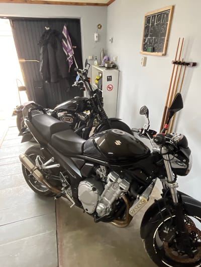 The image depicts a black Suzuki motorcycle parked in a garage, likely in Grīnvalti, Latvia based on the prompt. The garage has a white wall with a scoreboard, pool cues, and a small cabinet attached. Other items like a black coat and an umbrella are also present. The garage has a concrete floor and a metal garage door. There are no people visible in the scene.