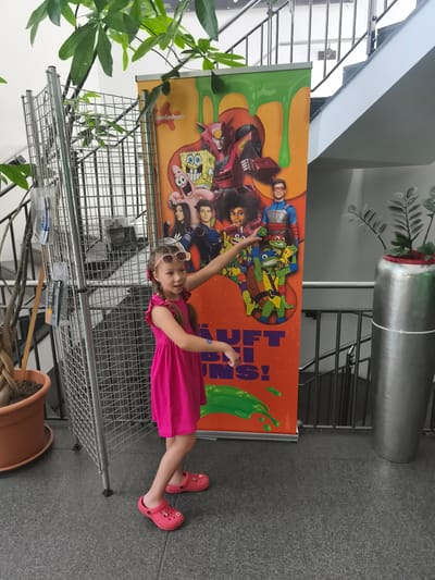 A young girl with blonde hair in pigtails, wearing pink sunglasses and a bright pink dress, stands to the left of a large, orange promotional banner. The banner features cartoon characters from Nickelodeon, including SpongeBob SquarePants, Teenage Mutant Ninja Turtles, and characters from "The Thundermans." The girl is pointing towards the banner with her right hand. She is wearing pink clogs. To the left of the girl is a metal display rack with various items attached to it. A large plant in a brown pot is also visible to the girl's left. Behind the banner, a staircase with metal railings is visible. The scene takes place indoors, on a grey tiled floor. The banner indicates that the event is in Niederkassel, Germany, as suggested by the text "Es läuft in Niederkassel!".