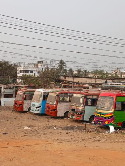 The scene depicts an unpaved, reddish-brown dirt lot in Boliarpur, Bangladesh, under a light gray, overcast sky. Six small to medium-sized buses or minibuses are parked in a linear formation, oriented towards the viewer's right.
From left to right: The leftmost vehicle, partially visible, appears as a stripped chassis or bus frame, adjacent to a large white rectangular object. The second bus features a red front and a light grey body. The third bus has a light blue front and a light grey body. The fourth bus displays an orange-red front and a light grey body with intricate detailing around the grille. The fifth bus has an orange-red front, a light blue body, and is extensively decorated on its front fascia; its side bears Bengali text, including "MAYER DUYA". The sixth and furthest right bus has a red and green body and a highly colorful, multi-hued front. Several buses exhibit roof racks.
In the foreground, a small, dark metallic stool or table stands on the dirt. Debris, including paper and plastic items, is scattered across the ground.
In the mid-ground and background, dense vegetation, including palm trees, partially obscures a collection of multi-story buildings, some with white and light blue facades, topped with water tanks. Overhead, numerous dark power lines traverse the upper portion of the frame. One individual is visible in the distant background, silhouetted atop a building structure. No direct interactions or actions are discernible among the visible people.