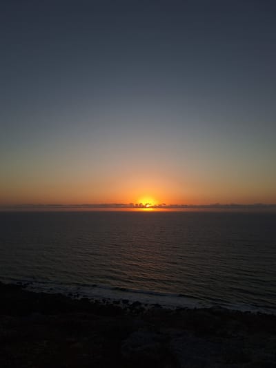 The image depicts a serene sunset over the ocean, likely near Ahrara, Morocco. The sun is a bright, glowing orb, partially obscured by a thin band of clouds at the horizon. Its warm, orange light casts a gentle glow across the water, creating shimmering reflections. The sea is calm, with gentle waves lapping at the shore. The foreground features a dark, rocky coastline, providing a stark contrast to the illuminated sky and sea.

The sky transitions from a deep, dusky blue at the top of the frame to a warm gradient of orange and yellow as it nears the horizon. This color palette evokes a sense of peace and tranquility. The overall mood is one of natural beauty and quiet contemplation. The time of day is clearly sunset, indicated by the low angle of the sun and the warm, diffused light. The presence of the ocean suggests a coastal location, and the rocky shore adds to the natural, untamed feel of the scene. There is no visible text in the image.