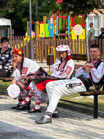 Four individuals are seated on a dark brown wooden bench outdoors. The two individuals on the left are women, and the two on the right are men. All four individuals appear to be of Bulgarian ethnicity and are dressed in traditional Bulgarian clothing. The woman on the far left is wearing a dark blue floral print top. The woman next to her is wearing a white blouse with red embroidery and a black skirt with red and white patterns. She has a red and yellow flower garland in her braided hair. She is holding a white circular object in her lap. The man in the center is wearing a white shirt with red embroidery and a black vest with intricate red and gold patterns. He has tattoos on his arms and is holding a water bottle. The man on the far right is wearing a white shirt with red embroidery and a black vest with red and gold trim. He has a white head covering with red embroidery. In the background, there is a colorful playground with red, yellow, and blue structures. A wooden fence separates the seating area from the playground. A sign with Bulgarian text is visible on the playground structure. The ground is paved with light gray interlocking bricks. Trees with green foliage are visible in the background, casting shadows. The overall scene suggests a cultural event or gathering in Pordim, Bulgaria.